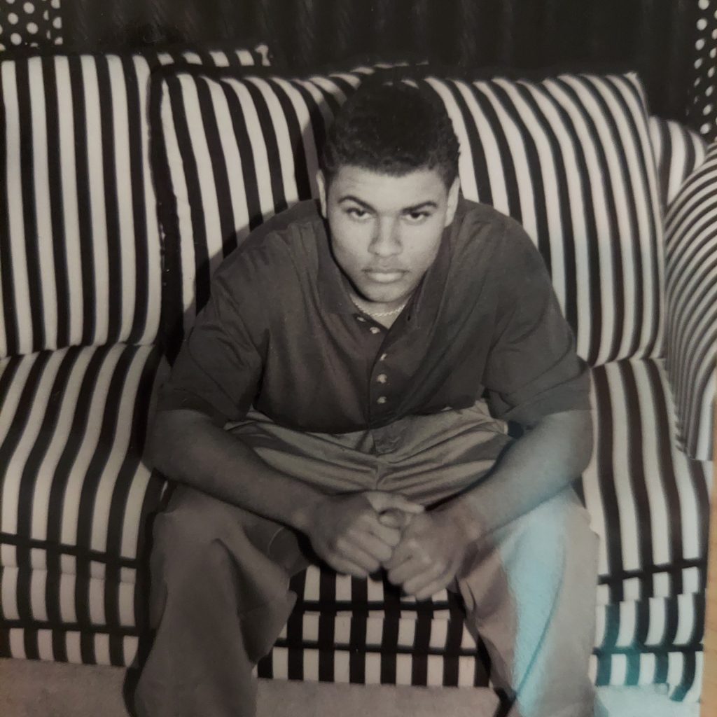 Black-and-white portrait of Andre Ramone seated on a striped couch, wearing a polo shirt and baggy pants, looking directly at the camera